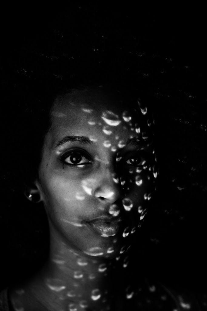 A woman's face half in shadow, the lit side dotted with projected water droplets.